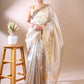 Woman wearing a beige Tissue saree with a wooden stool and plant in the background